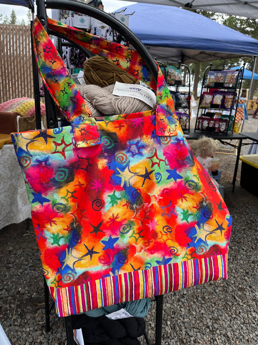 A multicolored tote bag with a vibrant swirl pattern, displayed on a stand at a market.